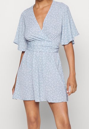 Kvinde iført en lyseblå, blomsterprintet omslagskjole med korte flagrende ærmer og dyb V-hals, stående mod en ensfarvet baggrund.