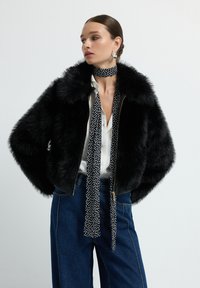 Czarna kurtka z sztucznego futra z wysokim kołnierzem, zapinana na zamek, z oversize'owymi rękawami, w zestawieniu z białą bluzką i chustą w kropki oraz niebieskimi, szerokimi spodniami dżinsowymi.