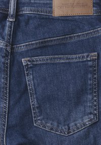 Denim w ciemnoniebieskim kolorze o teksturowym wykończeniu. Posiada jedną tylnią kieszeń oraz naszywkę z brązowej skóry z logo marki wszytą w pasie.