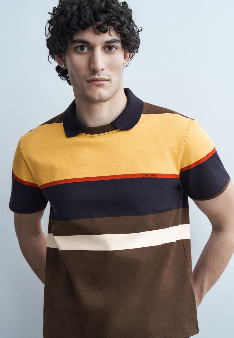 Young man with curly hair wearing a multicolored polo shirt with brown, yellow, navy, and white horizontal stripes, standing against a plain light background.