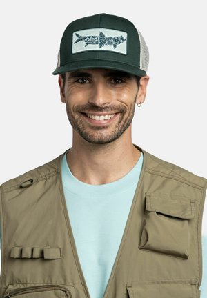 Lächelnder Mann mit einer grünen Kappe mit Fischmotiv, hellblauem Hemd und beiger Utility-Weste mit mehreren Taschen.