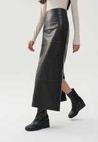Černá kožená maxi sukně s hladkým povrchem, se švy a bočním rozparkem, doplněná o elegantní černé kotníkové boty na wedge podpatku.