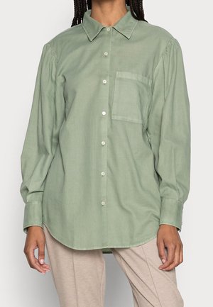 Femme portant une chemise ample verte claire à boutons avec de longues manches bouffantes et un pantalon beige, debout devant un fond uni.
