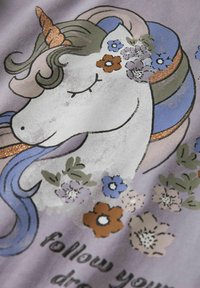 Paarse stof met een eenhoornontwerp met een gouden hoorn, kleurrijke manen en bloemrijke accenten. Tekst luidt "volg je dromen."