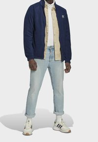 Marinblå fleecejacka, beige skjorta med knappar, ljusblå jeans och vita sneakers med svarta ränder och bruna sulor. Avslappnad, lager-på-lager-outfit.