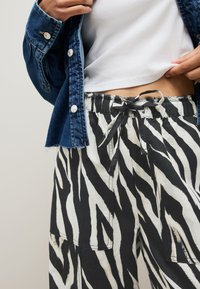 Person wearing black and white zebra-striped pants with a drawstring waist, a white ribbed top, and a denim jacket with metal buttons.