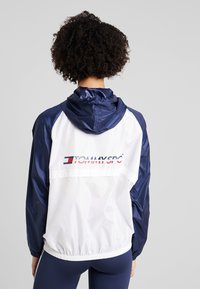 Giacca a vento bianca e blu navy con cappuccio, orlo elastico e logo sul retro. Materiale liscio e leggero sembra resistente all'acqua.