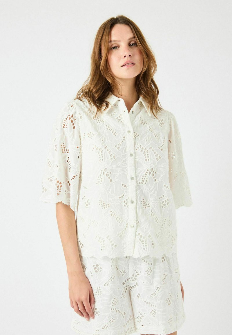 Hvit brodert skjorte med kort erme, preget av intrikate blomsterdesign og bølgete kanter, parret med matchende shorts.