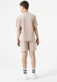 Conjunto de toalla de color marrón claro, con camiseta de manga corta y pantalones cortos. Llevado con zapatillas blancas y calcetines con detalles en negro. Diseño simple.
