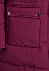Liela dziļi purpura quilted jaka ar kabatu, kurai ir vāks, ko noslēdz divi sudraba pogas.