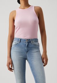 Mujer vestida con una camiseta de tirantes rosa de canalé y unos vaqueros de tiro alto azul claro, frente a un fondo liso.