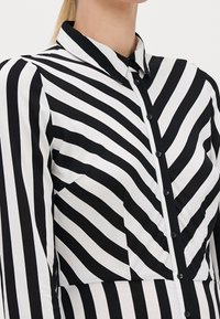 Zbliżenie na osobę noszącą czarno-białą koszulę z guzikiem w pionowe i ukośne paski, z kołnierzykiem i długimi rękawami.