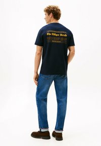 Joven de pie, vistiendo una camiseta gráfica azul marino con texto amarillo y jeans azules enrollados en los puños, zapatos marrones sobre un fondo blanco.