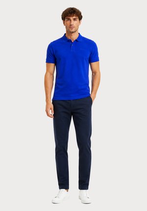 Homme portant un polo bleu vif à manches courtes, un pantalon bleu marine foncé et des baskets blanches, debout avec une main dans la poche.