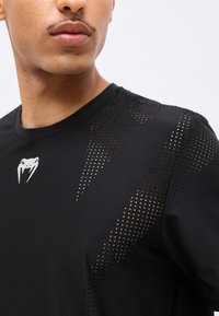 Κοντινό πλάνο ενός ατόμου που φοράει μαύρο διάτρητο αθλητικό μπλουζάκι με λευκό λογότυπο κυνόδοντα φιδιού στο στήθος.