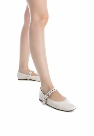 Zapatos planos blancos con puntera redondeada, con una correa adornada con pequeños remaches metálicos, exhibidos sobre un fondo liso.