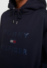 Sudadera navy con cuello ajustable. Presenta un logo brillante "TOMMY HILFIGER" y un pequeño detalle de bandera tricolor en el centro. Textura suave.