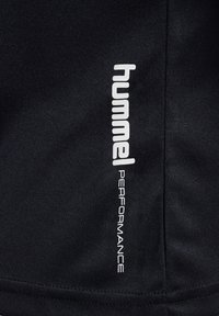 Sort sportswear stof med et hvidt trykt logo "hummel" og "PERFORMANCE" lodret langs sidesømmen. Glat tekstur og mat finish.