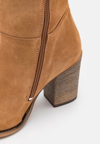 Beżowy zamszowy botek za kostkę z teksturowanym, drewnianym obcasem, bocznym zamkiem błyskawicznym i gładkim wykończeniem. Charakteryzuje się zaokrąglonym noskiem i delikatnymi przeszyciami.