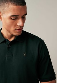 Polo de color verde oscuro hecho de tejido texturizado, con cuello, dos botones y un pequeño emblema de un ciervo dorado en el pecho.