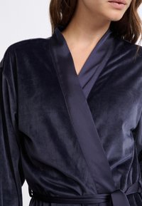 Femme portant un peignoir en velours bleu marine avec un col et une ceinture en satin, montrée des épaules à la taille sur un fond uni.