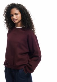 Sudadera burdeos con cuello redondo y puños acanalados. Presenta un corte holgado y una tela texturizada. Combinada con pantalones azul oscuro.