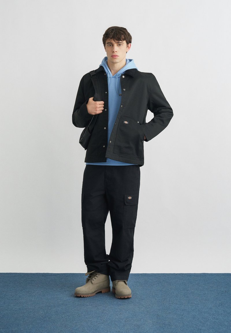 Giovane uomo con giacca nera sopra una felpa con cappuccio blu, pantaloni cargo neri, stivali beige e una borsa a tracolla nera.