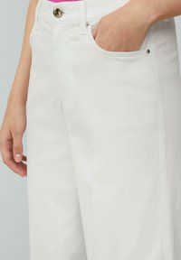 Pantalon en coton blanc avec une texture lisse, doté d'une poche avant et d'une fermeture à bouton en métal. Couture visible le long des coutures.
