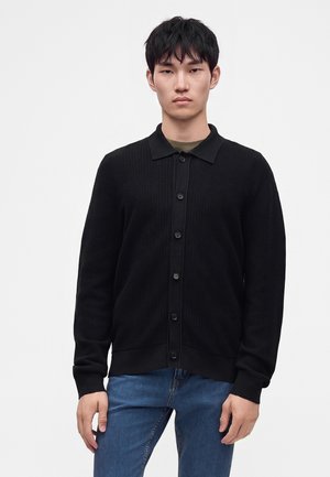 Jeune homme portant un cardigan noir boutonné et un jean bleu, debout devant un fond blanc uni.