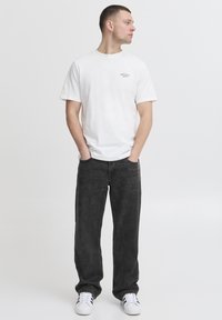 Camiseta blanca de algodón con un pequeño logo negro en el pecho izquierdo, combinada con jeans holgados de color gris oscuro y zapatillas blancas.