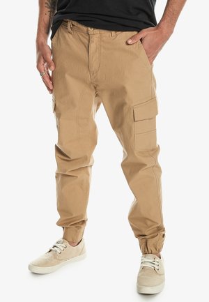 Pantalones cargo beige con puños elásticos, bolsillos laterales y un corte relajado. Hechos de un tejido ligero con detalles de costura visibles.