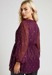 Blusa plumetis trasparente in tonalità prugna con motivi argentati simili a piume, vita arricciata e maniche a sbuffo. Texture liscia con design in tessuti sovrapposti.
