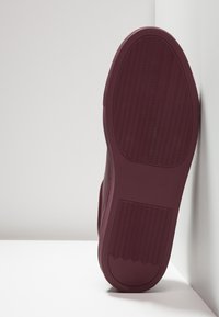 Maroon sneaker with a textured rubber sole, featuring a wavy pattern, rounded shape, and a sleek design aligned against a white wall.