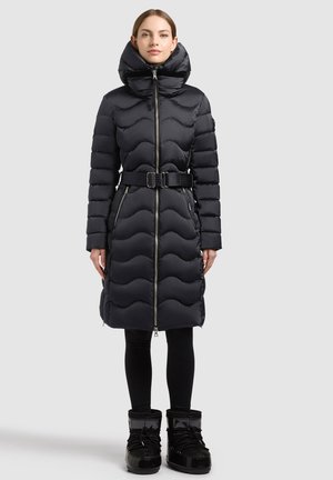 Frau trägt einen langen schwarzen gesteppten Steppmantel mit hohem Kragen, Gürtel, Frontreißverschluss, schwarze Leggings und schwarze Winterstiefel, stehend.
