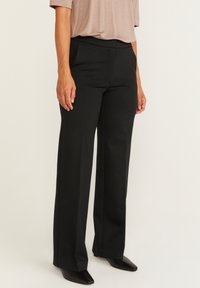 Zwarte rechttoe rechtaan broek met een gladde textuur en een brede tailleband. Gepaard met puntige zwarte schoenen. Draag met een lichtbruine top.