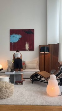 Scena w salonie z beżową sofą, marmurowym stolikiem kawowym, jasnymi drewnianymi podłogami, abstrakcyjnymi dziełami sztuki na ścianie i białą okrągłą lampą. Dywan o teksturowanym futrze.