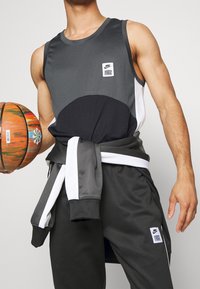Le débardeur de sport Nike présente une palette de couleurs gris et noir avec une texture en maille, un patch logo, associé à un pantalon assorti et un ballon de basket.