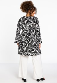 Schwarz-weiße, blumengemusterte Tunika mit langen Ärmeln und Seitenenschlitzen, kombiniert mit weißen, weit geschnittenen Hosen und schwarzen Sandalen mit Blockabsatz.