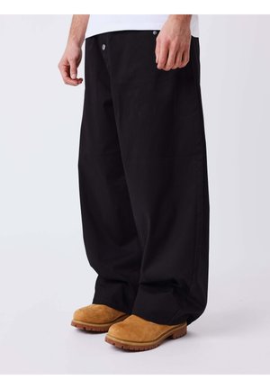 Personne portant un pantalon noir ample et des bottes de travail en daim beige debout sur un fond blanc.