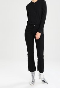 Persona che indossa un top nero a maniche lunghe con bottoni, pantaloni attillati neri e stivaletti con tacco a blocco argentati brillanti, in piedi su uno sfondo bianco.