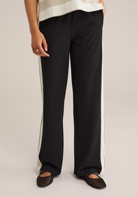 Pantalons larges noirs avec des bandes latérales blanches, fabriqués en tissu lisse. Le modèle porte des ballerines en mesh noires, se tenant sur un fond neutre.