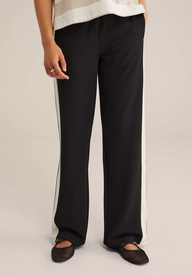Pantalons larges noirs avec des bandes latérales blanches, fabriqués en tissu lisse. Le modèle porte des ballerines en mesh noires, se tenant sur un fond neutre.