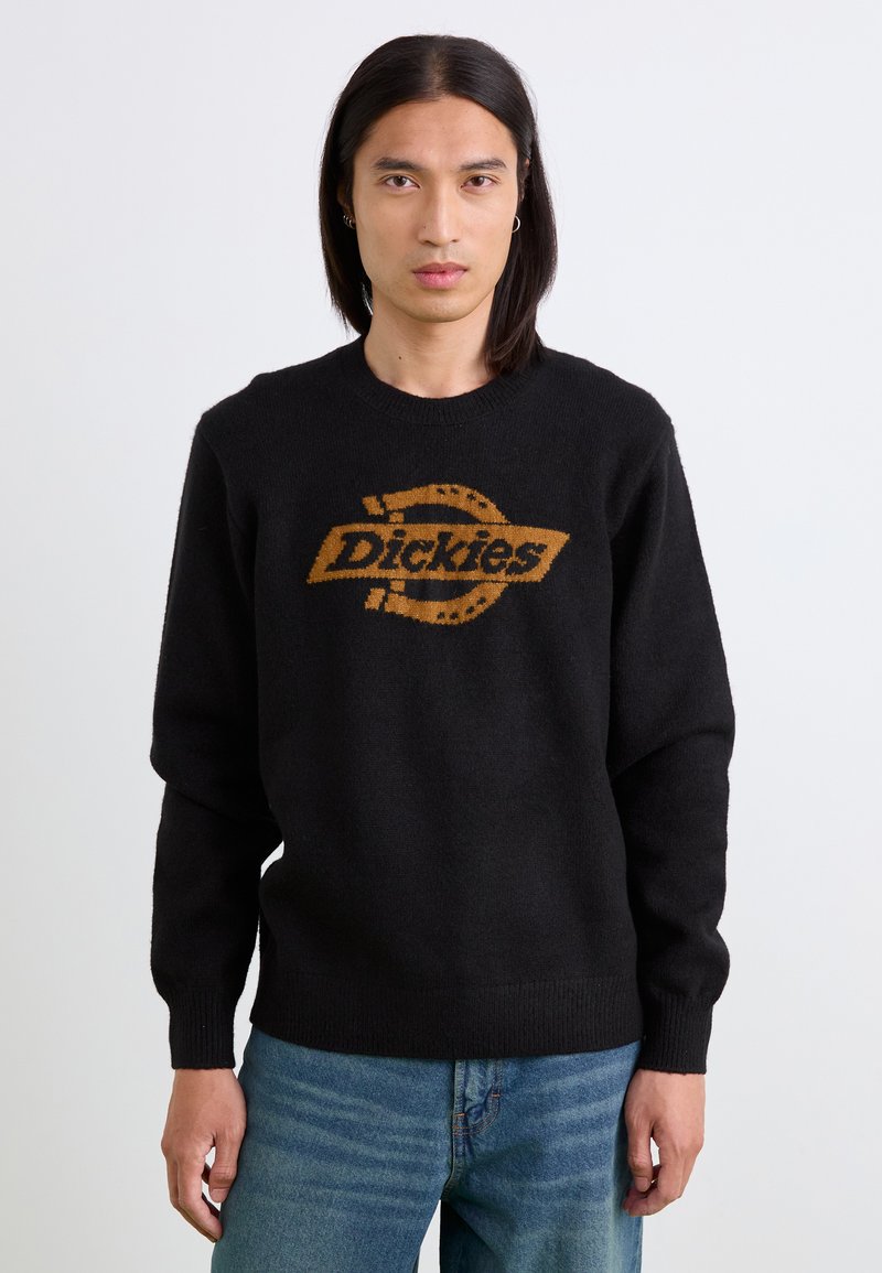 Schwarzer Strickpullover mit einem kontrastierenden braunen "Dickies"-Logo, rundem Halsausschnitt und gerippten Bündchen sowie Saum. Getragen mit blauen Jeans.