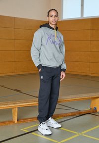 Sudadera gris con diseño geométrico púrpura, acentos negros y un cordón amarillo, combinada con pantalones de chándal negros y zapatillas blancas.