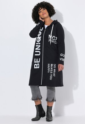 Black zip-up hooded long jacket with white text graphics, paired with grey cropped jeans featuring frayed hems and black studded ankle boots.