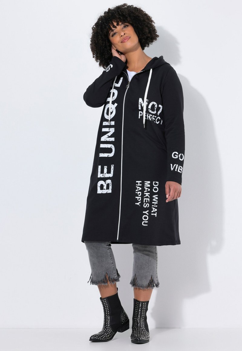Zwarte ritssluiting capuchon lange jas met witte tekstgraphics, gecombineerd met grijze cropped jeans met rafelige zomen en zwarte gespijkerde enkellaarsjes.