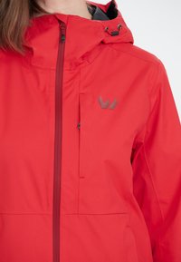 Veste imperméable rouge avec une fermeture éclair à l'avant, une capuche et un accent logo gris. Présente une texture lisse et un design contemporain élégant.
