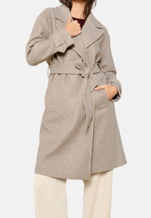 Kvinde iført beige uldfrakke med bælte, brede revers og lommer, kombineret med cremefarvede ribstrikkede bukser.