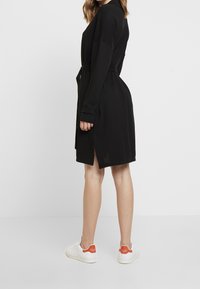 Robe noire ceinturée avec de longues manches, dotée d' fentes sur les côtés et d'une coupe décontractée, associée à des baskets blanches avec des accents orange.