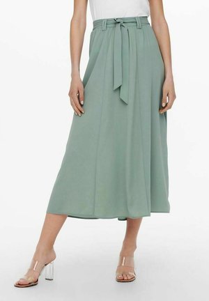 Femme portant une jupe midi taille haute vert sauge avec une ceinture nouée et des sandales transparentes à talons, debout avec une jambe légèrement en avant.